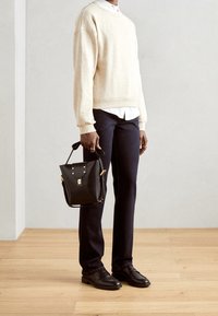 Suéter beige, camisa blanca, pantalones oscuros y zapatos de cuero negros. Sosteniendo un bolso de cuero negro con herrajes dorados y un diseño estructurado.