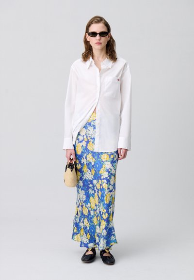 Chemise blanche à boutons, jupe maxi florale bleue et jaune, chaussures noires et petit sac en osier au design arrondi.