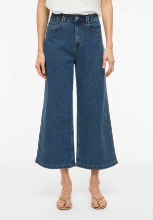 Sieviete valkā augsta vidukļa, platu kāju zilu džinsa bikses, kas saīsinātas virs potītēm, kopā ar bēša krāsas sandales ar siksniņu un baltu iešūtu topu.