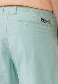Shorts en teal clair en tissu doux, avec une poche arrière et une étiquette noire cousue sur la taille avec "S.O. Elevated Casual, Est. 1969".