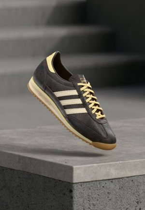 Ciemnobrązowy sneaker Adidas z kremowymi paskami i sznurowadłami, beżowym akcentem na pięcie i gumową podeszwą, umieszczony nad betonową powierzchnią.
