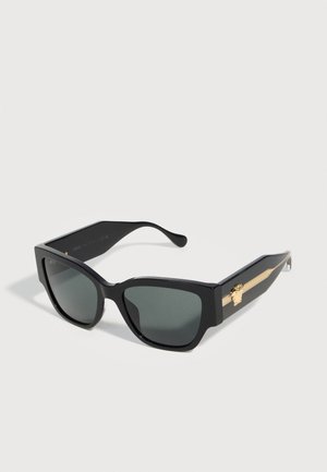 Lunettes de soleil rectangulaires noires oversize avec des verres teintés sombres et des accents décoratifs dorés sur les branches.