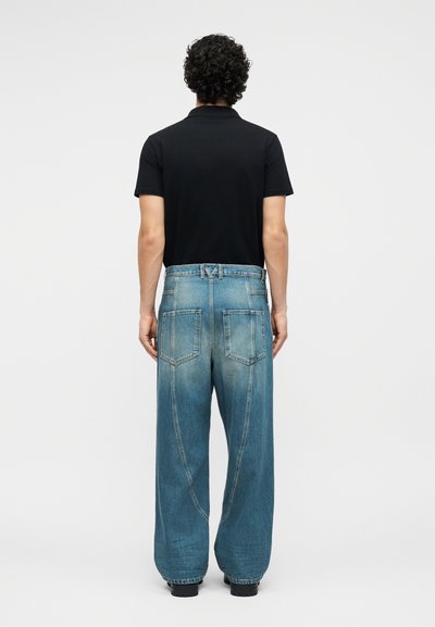 Άνδρας με σγουρά μαλλιά, φορόντας μαύρο κοντομάνικο πουκάμισο και φαρδιά μπλε τζιν, στέκεται πλάτη σε μονόχρωμο φόντο.