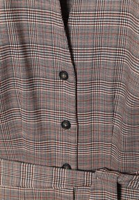 Abito a quadri con una combinazione di colori marrone, nero e bianco. Presenta un profondo scollo a V, cinque bottoni e una cintura coordinata per accentuare il punto vita.