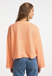 Pull léger en tricot gaufré à manches longues de couleur orange pastel doux. Il présente un col rond et une coupe décontractée. Associé à un jean en denim taille haute.