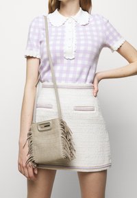 Kobieta w topie w lilacowe i białe kratki z krótkim rękawem, białej mini spódniczce z tweedu oraz beżowej torebce crossbody z frędzlami.