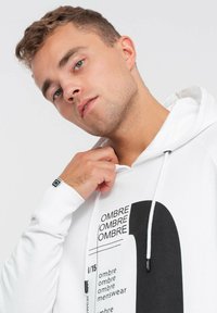 Sudadera blanca con un gran diseño gráfico negro acompañado de texto. Cuenta con cordones ajustables y un logo en la manga. Textura suave, corte casual.