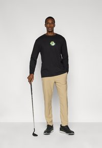 Μαύρο μακρυμάνικο πουκάμισο με πράσινο γραφικό, μπεζ παντελόνια και μαύρα παπούτσια γκολφ. Το μοντέλο κρατάει ένα γκολφ κλαμπ.