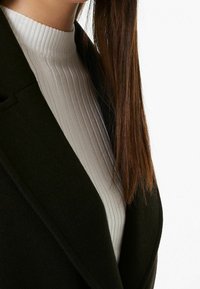 Černý kabát z vlněné směsi s šálovým límcem, vrstvený přes světle šedý žebrovaný top s vysokým výstřihem, který má hladkou texturu a slim fit.