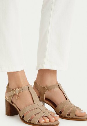 Béžové semišové sandály na podpatku s kotníkovými pásky, zlatou přezkou a dekorativním přívěskem, nošené s bílými zkrácenými kalhotami.