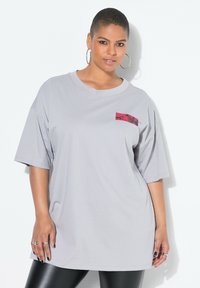 Světlé šedé oversized tričko z měkkého materiálu, s červeným potiskem na levém hrudním prostoru. Krátké rukávy a volný střih.