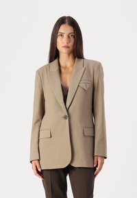Blazer beige ajusté, fermeture à un bouton, deux poches avant et un détail en triangle sur la poitrine. Tissu lisse et structuré.