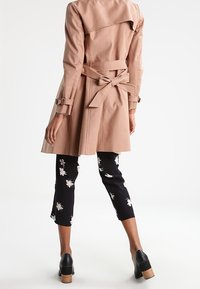 Beżowy płaszcz trench z paskiem w talii, długimi rękawami i wentylacją z tyłu; w połączeniu z czarnymi kwiecistymi spodniami typu cropped oraz czarnymi butami na słupku.
