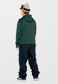 Zielona bluza z kapturem w połączeniu z granatowymi spodniami cargo. Akcesoria to beanie w kolorze piaskowym oraz gogle narciarskie. Czarno-białe sneakersy.