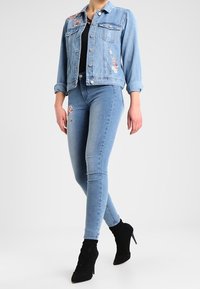 Chaqueta de mezclilla azul claro con bordado floral, combinada con jeans ajustados azul oscuro y botines negros de tacón alto.