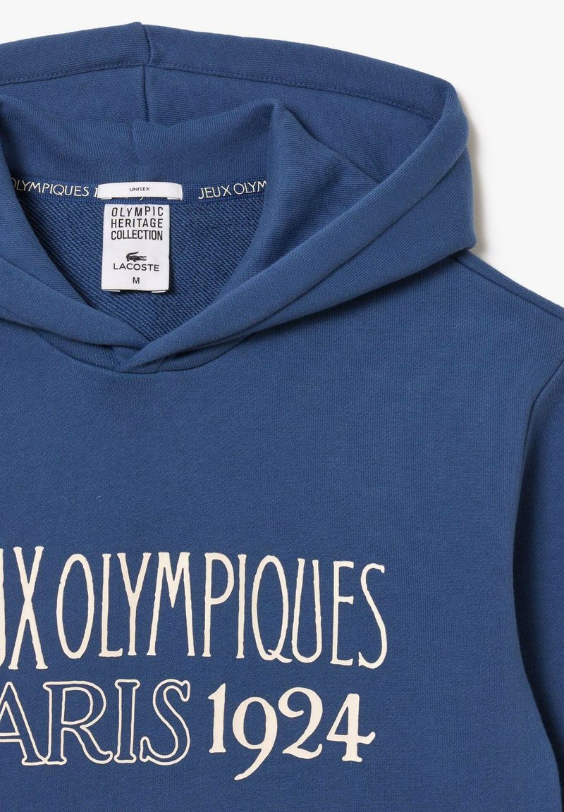 Blauer Kapuzenpullover mit dem Aufdruck "Jeux Olympiques Paris 1924" in Weiß und einem Etikett mit der Aufschrift "Olympic Heritage Collection Lacoste", Größe M.
