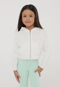 Ragazza giovane in piedi con una felpa bianca con zip e leggings verde chiaro su uno sfondo bianco uniforme.