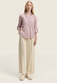Chemise rose clair texturée à boutons avec manches retroussées, associée à un pantalon large crème et des chaussures mule dans une teinte douce blanc cassé.