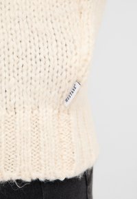 Sweter w kolorze kremowym z dzianiny, z prążkowanym wykończeniem i małą białą metką z napisem "MUSTANG". Miękka faktura z grubą nitką.