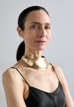 Zlatý kovový náhrdelník ve tvaru obojku s vlnitým vzorem, nošený přes černý top bez rukávů s tenkými ramínky a detailem knoflíku.