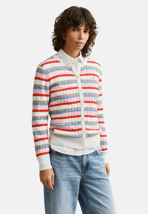 Femme portant un pull blanc boutonné avec des rayures horizontales rouges, bleues, beiges et roses, par-dessus une chemise blanche à col et un jean bleu clair.