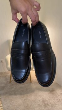 Main tenant une paire de mocassins slip-on en cuir noir avec des semelles intérieures "PIER·ONE", à l'intérieur sur un sol recouvert de moquette près d'une chaise.