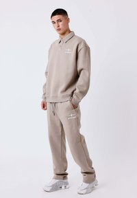 Beige sweatshirt with collar and logo on chest, paired with matching joggers featuring pockets and logo. White sneakers complete the look.
