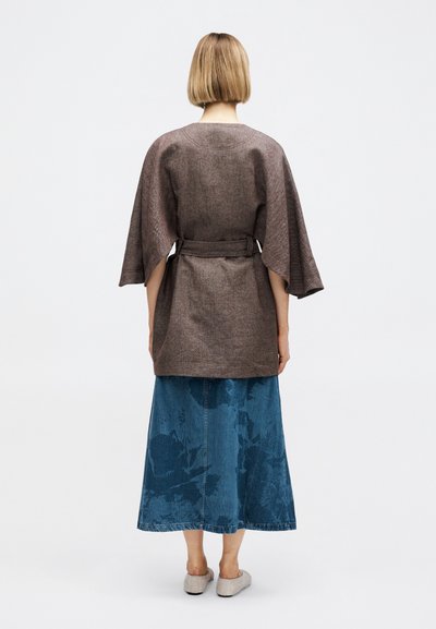 Άτομο που στέκεται με την πλάτη γυρισμένη, φοράει καφέ μπουφάν με ζώνη, μπλε φούστα με σχέδια και μπεζ επίπεδα παπούτσια σε λευκό φόντο.