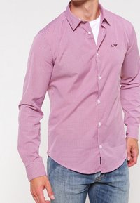 Muž v úzké košili s dlouhými rukávy v růžovém kostkovaném vzoru přes bílém tričku, v modrých džínech, stojící s jednou rukou u boku.