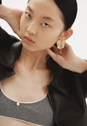 Pendientes texturizados de oro con un detalle de caída, un delicado collar de cadena de oro con un colgante cuadrado, superpuestos sobre un top de punto gris y una chaqueta negra.