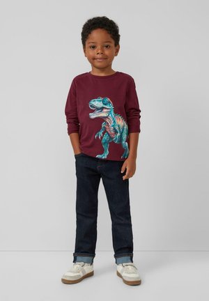 Bordeaux langarm overhemd met een grafiek van een kleurrijke T-Rex, gecombineerd met donkerdenim jeans en witte sneakers met beige accenten.
