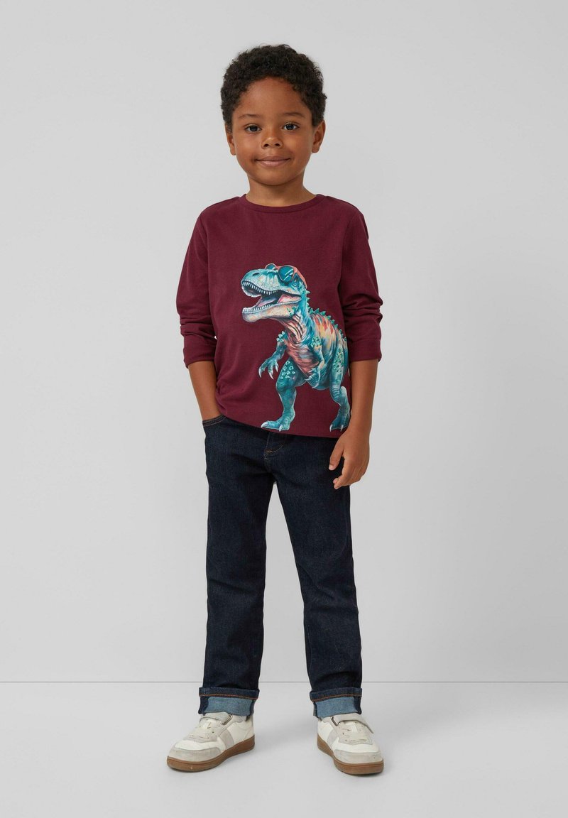 Burgunderfarbenes Langarmhemd mit einem Grafikdruck eines bunten T-Rex, kombiniert mit dunkelblauen Jeans und weißen Sneakers mit beigen Akzenten.