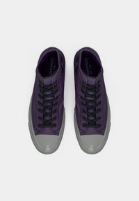 Violet krāsas auduma sporta apavi ar melnajām auklām, pelēkiem gumijas pirkstgaliem un gludu virsmu. Uz mēles redzama Converse zīmola atzīme.