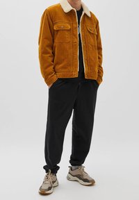 Giacca di velluto a costine gialla senape con collo in shearling; due tasche sul petto; abbinata a pantaloni jogger neri e sneakers beige.
