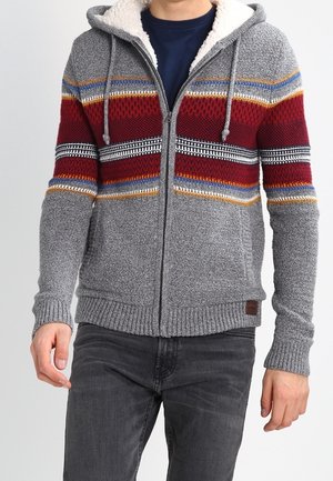 Mies, jolla on harmaa neulottu vetoketjullinen huppari, jossa on punaisia, sinisiä, valkoisia ja sinapinvärisiä vaakaraitoja, yhdistettynä tummanharmaisiin farkkuihin.