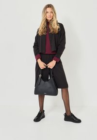 Svart zip-up-jacka över en burgundy ribbad topp, tillsammans med svarta shorts och sneakers. En strukturerad svart handväska hålls. Snygg hudton.