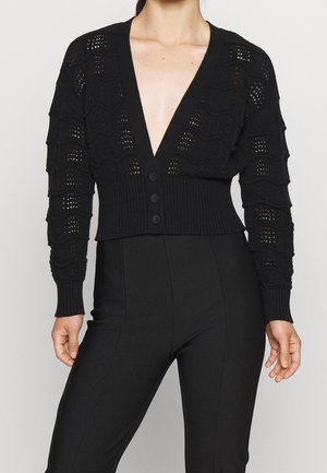 Black cropped cardigan with textured wave patterns, ribbed waistband, and three front buttons, worn with high-waisted fitted black pants.