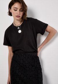 T-shirt in cotone nero con maniche arrotolate abbinato a una gonna nera testurizzata con accenti circolari, il tutto su uno sfondo neutro.