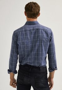 Chemise à manches longues bleu foncé avec motif quadrillé blanc. Dotée d'un col plié et de poignets boutonnés. Portée avec un jean noir.