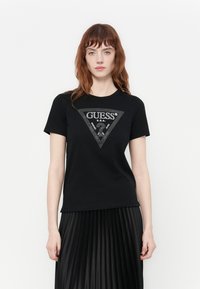 Black cotton t-shirt featuring a triangular logo design with rhinestones. Short sleeves and crew neckline. Worn with a pleated black skirt.