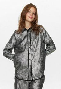 Camicia metallica argentata con maniche lunghe, caratterizzata da un bordo nero lungo i bottoni e il colletto, che mostra un tessuto liscio e strutturato.