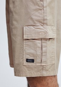 Beige cargoshorts i letvægtsstof med en stor sidelomme med klap, syningsdetaljer og en etiket med tekst.