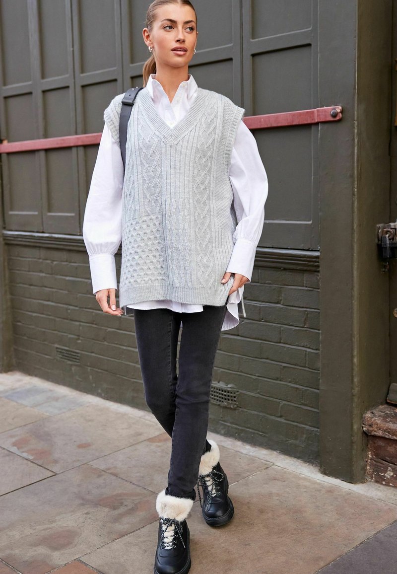 Gilet en tricot gris avec des motifs en câble et texturés sur une chemise blanche, associé à un jean noir slim-fit et des bottes noires avec des accents en fourrure.