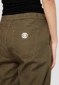 Olijfgroene katoenen broek met een elastische tailleband, voorzien van een enkele achterzak versierd met een zilveren logo detail.
