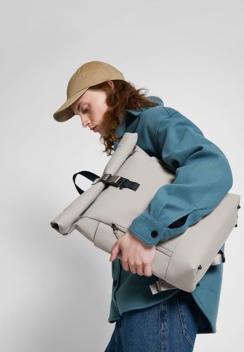 Rucsac gri cu clapetă, cu o cataramă neagră și curea ajustabilă, ținut lângă o geacă teal, având o textură netedă și un design minimalist.