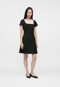 Γυναίκα με μακριά σκούρα μαλλιά που φοράει ένα μαύρο κοντομάνικο εφαρμοστό φόρεμα, λευκή σειρά από μαργαρίτες και μαύρες μυτερές φλατ, που στέκεται σε άσπρο φόντο.