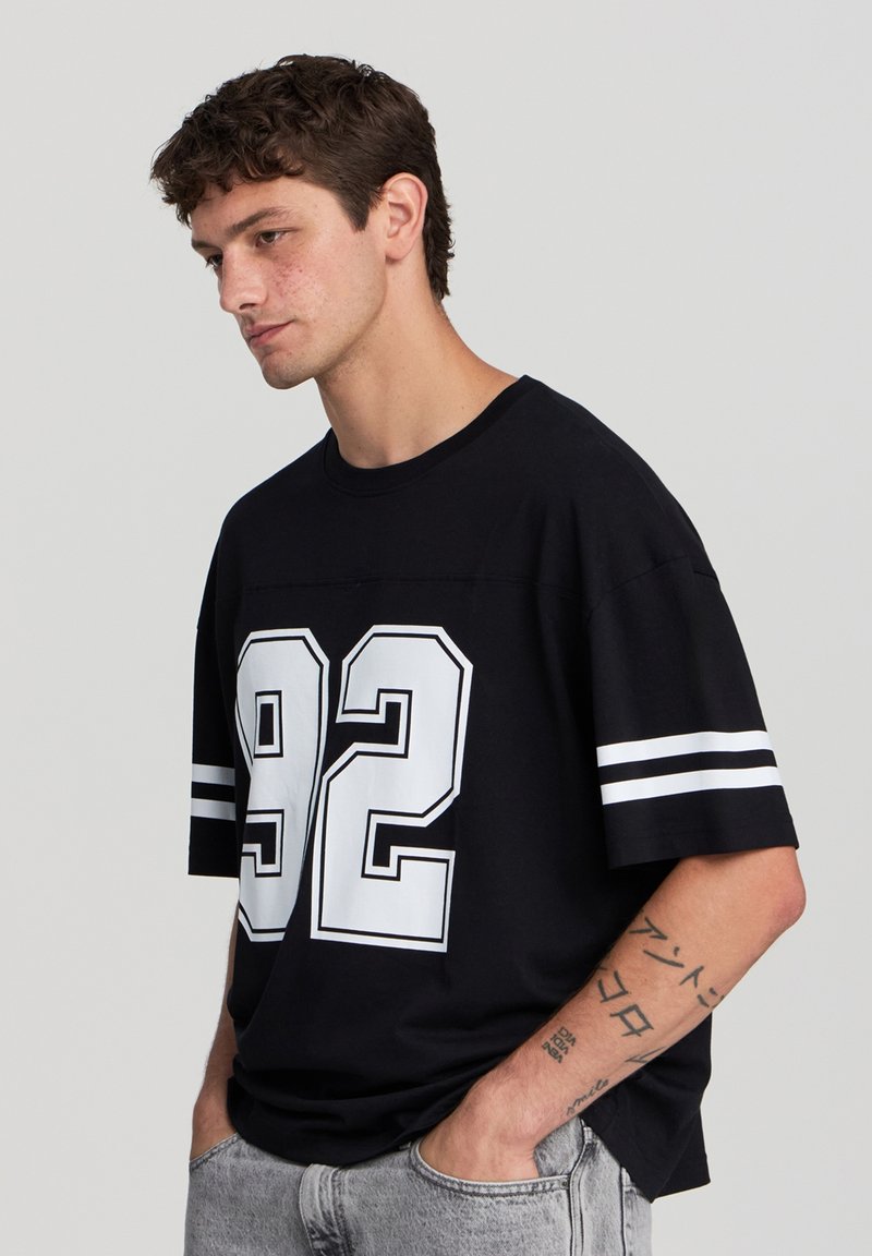 Czarna, oversize koszulka z białym nadrukiem "92" i paskowanymi rękawami. Materiał wydaje się miękki; okrągły dekolt. Model stoi bokiem.