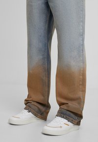 Plačios kojos džinsai su gradientiniu išblukimu nuo šviesiai mėlynos iki rudos, dėvimi su baltais sportbačiais, turinčiais smėlio spalvos akcentų.