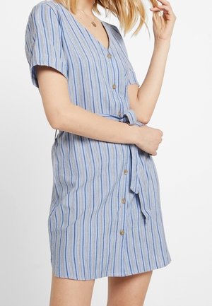 Femme portant une robe bleue à rayures, à manches courtes, boutonnée à l'avant, avec une ceinture en tissu nouée à la taille, sur un fond blanc.