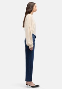 Maglione beige oversize con maniche a sbuffo, abbinato a jeans blu scuro a vita alta e décolleté nere con punta appuntita. Vista di profilo laterale.